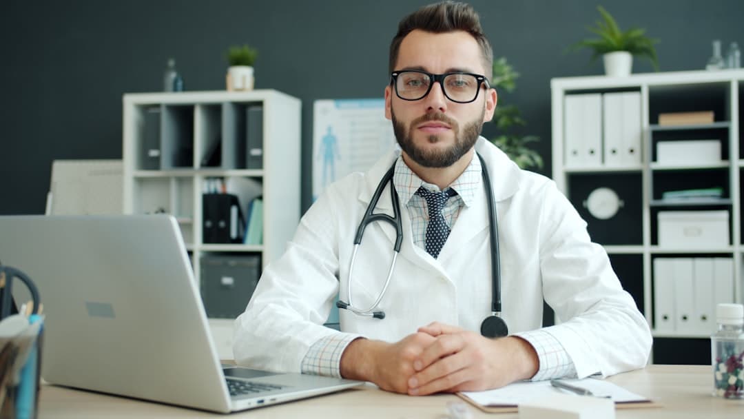 Doctor wearing a white coat sits at a desk.. Cover image for: When AI Scribes Slow You Down: The First Two Weeks Slump and How to Push Through.