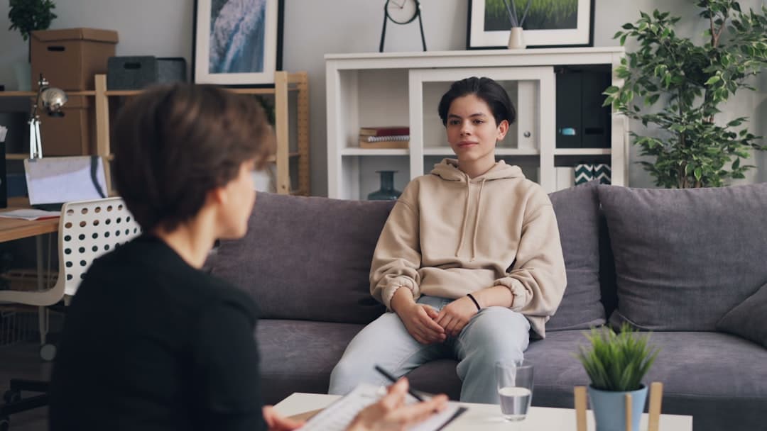 a woman sitting on a couch talking to another woman. Cover image for: AI Scribe for Psychiatry and Mental Health: What's Different.