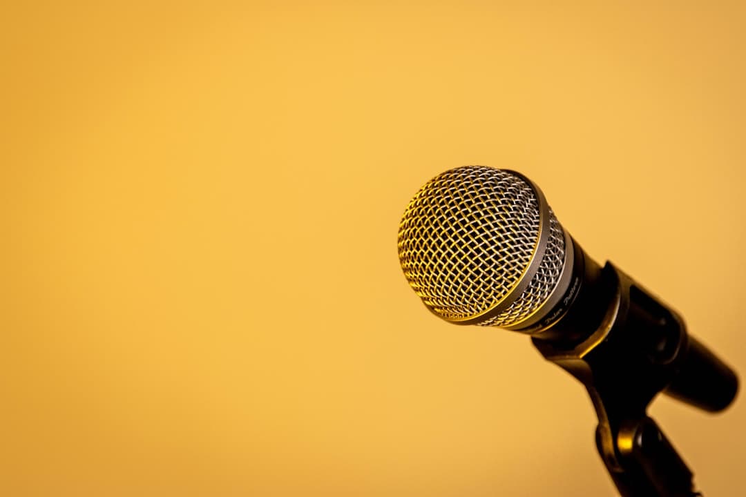 A microphone on a stand against a yellow background.. Cover image for: From Ambient to Agentic: How AI Scribes Are Starting to Do More Than Document.